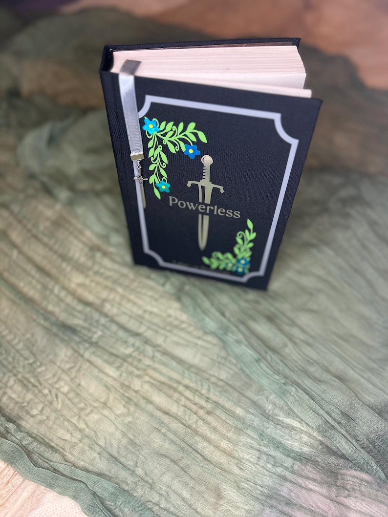 May include: A hardcover book with a black cover featuring the word "Powerless" and a sword illustration. The cover is decorated with floral and leaf designs in blue and green. A silver ribbon bookmark is attached.