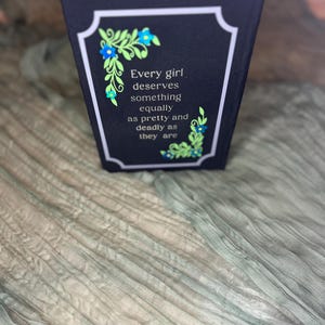 May include: A black book cover with a white border and the quote "Every girl deserves something equally as pretty and deadly as they are." Floral accents in blue and green adorn the corners. The book rests on a textured, light green fabric.