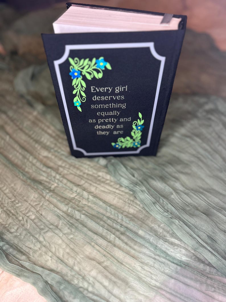 May include: A black book cover with a white border and the quote "Every girl deserves something equally as pretty and deadly as they are." Floral accents in blue and green adorn the corners. The book rests on a textured, light green fabric.
