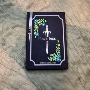 May include: A hardcover book titled "Powerless" by Lauren Roberts. The cover is black with a silver sword illustration and floral accents in green and blue. A silver ribbon and sword charm are attached.