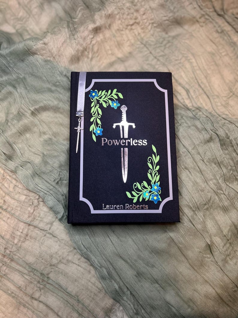 May include: A hardcover book titled "Powerless" by Lauren Roberts. The cover is black with a silver sword illustration and floral accents in green and blue. A silver ribbon and sword charm are attached.