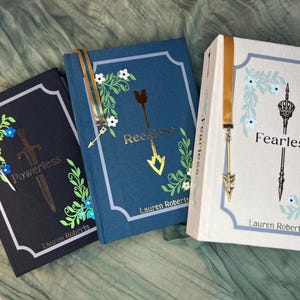 May include: Three hardcover books with floral and arrow designs. The books are titled "Powerless", "Reckless", and "Fearless". Each book has a decorative bookmark and the author's name, Lauren Roberts, is printed on the bottom.