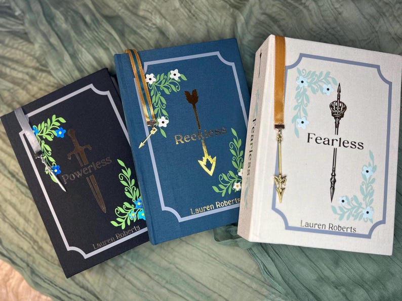 May include: Three hardcover books with floral and arrow designs. The books are titled "Powerless", "Reckless", and "Fearless". Each book has a decorative bookmark and the author's name, Lauren Roberts, is printed on the bottom.
