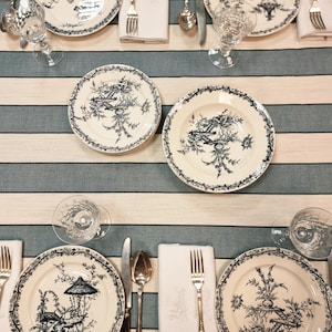 May include: Overhead shot of a table setting with six plates featuring a blue and white landscape design. Each place setting includes silverware, a folded white napkin, and a water glass. The tablecloth has blue and white stripes.