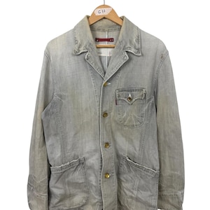 May include: A light grey denim jacket with a button-up front, two front pockets, and a small pocket on the chest. The jacket has a classic collar and is hanging on a wooden hanger. The jacket has a worn, vintage look.