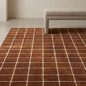 Pode incluir: Um tapete retangular castanho com um padrão de grade de blocos retangulares delineados em branco. Um banco de tons neutros é parcialmente visível no fundo. A textura do tapete sugere uma sensação suave e confortável.