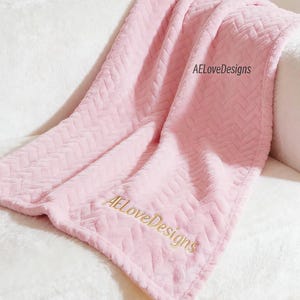 Könnte beinhalten: Eine weiche, hellrosa Wohndecke mit einem erhabenen Fischgrätmuster. Der Stoff hat eine flauschige, pelzige Optik. Der Schriftzug AELoveDesigns ist an zwei Stellen auf der Oberfläche der Decke mit goldfarbenem Garn eingestickt.