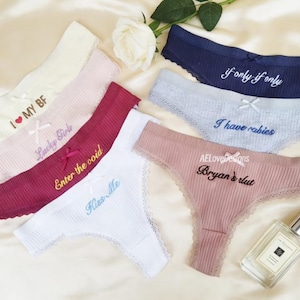May include: A collection of thong underwear in shades like cream, pink, burgundy, white, navy, and light blue. Each piece has embroidered text, including phrases like "I MY BF", "Lucky Girls", and "If only if only". A perfume bottle and a rose are also visible.