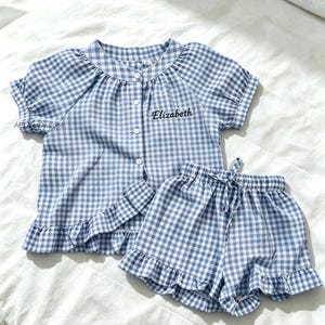 Könnte beinhalten: Ein blau-weiß kariertes Pyjama-Set. Das Set besteht aus einem kurzärmeligen Hemd mit Knöpfen und Rüschensaum sowie passenden Shorts mit Kordelzug und Rüschensaum. Der Name "Elizabeth" ist auf das Hemd gestickt.