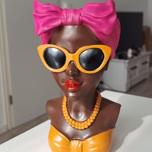 May include: A decorative bust featuring a woman with dark skin, wearing orange sunglasses, a pink headscarf, and an orange top. A yellow beaded necklace and red lipstick complete the look. The bust is a unique home decor item.