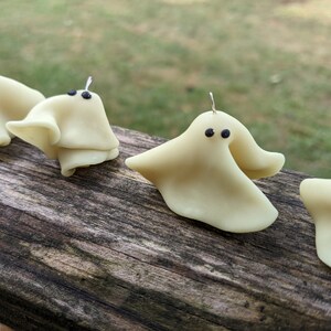 May include: Four small white ghost ornaments with black eyes. The ghosts are hanging from a wooden surface.