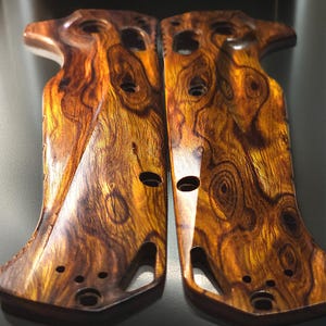 May include: Two wooden pistol grips with a rich, swirling grain pattern in shades of brown and gold. Each grip has a series of drilled holes and a contoured shape for comfortable handling. The grips are designed for a firearm and are made of a durable wood.