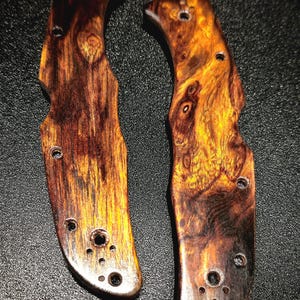 May include: Two knife handles with a natural wood grain pattern. The wood features a mix of dark brown, reddish-brown, and golden-yellow hues. Each handle has several drilled holes for attachment.