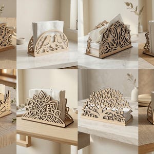 May include: A selection of laser-cut wooden napkin holders. Designs include butterflies, birds, and trees. Each holder contains white napkins and is displayed on a table. The holders are made of light-coloured wood.