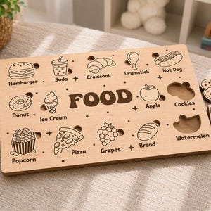 Può includere: Puzzle in legno a tema cibo, con sagome per vari alimenti come un croissant, una mela e dell'uva. La parola "FOOD" è ben visibile. Include pezzi rimovibili a forma di cibo.
