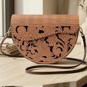 May include: A semi-circular handbag featuring a light brown wooden exterior with intricate laser-cut floral patterns and a solid black interior lining. The bag includes a thin brown leather shoulder strap attached with gold-toned metal hardware.