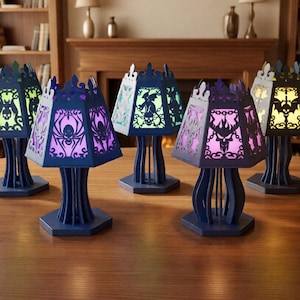 Può includere: Cinque lampade blu scuro con intricati disegni ritagliati raffiguranti ragni, teschi e pipistrelli. Ogni lampada ha una base esagonale e un paralume esagonale abbinato che emette una luce colorata, inclusi verde, viola, blu e giallo.
