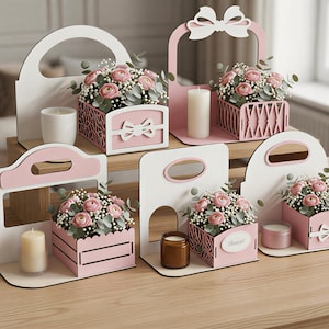 Könnte beinhalten: Eine Sammlung dekorativer Blumenkästen und Kerzenhalter aus Holz in Weiß und Rosa. Jede Box enthält rosa Rosen und Grünzeug. Einige haben Schleifen, andere Griffe. Kerzen in verschiedenen Größen sind ebenfalls vorhanden.