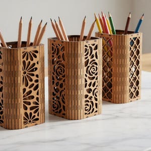 Könnte beinhalten: Drei Stifthalter aus Holz mit dekorativen Ausschnitten. Die Halter sind mit Bleistiften und Stiften in verschiedenen Farben gefüllt. Die Halter sind aus hellbraunem Holz gefertigt und stehen auf einer weißen Marmoroberfläche.
