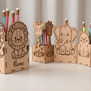 Op de afbeelding: Houten potlodenhouders met dierenontwerpen, waaronder een leeuw, een giraf, een olifant en een beer. Elke houder is lichtbruin en heeft een uitgesneden dierenfiguur. De potlodenhouders zijn gevuld met potloden in verschillende kleuren.
