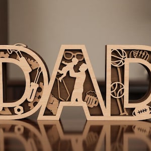 以下が含まれることがあります： 「DAD」という文字が切り抜かれたライトブラウンの木製看板。文字には、ハンマー、レンチ、ドライバーなどの工具、子供を持ち上げる人のシルエット、さまざまなスポーツ用品など、レーザーカットによる複雑なデザインが施されています。