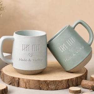 Puede incluir: Dos tazas de cerámica con acabado mate, una de color azul claro y otra verde salvia. Cada taza presenta un diseño de marcas de conteo grabado, un pequeño icono de corazón y el texto "Hale & Vicky" en letra cursiva con un asa lateral.