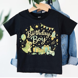 May include: Black short-sleeved t-shirt with a colorful graphic design. The design includes the text "Birthday Boy" with a dinosaur, gifts, a balloon, a rocket, and a game controller. The shirt is hanging on a wooden hanger.