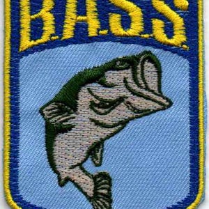 May include: A shield-shaped embroidered patch featuring the text B.A.S.S. in bold yellow thread at the top. Below the text, a light blue background displays a detailed, dark green and light gray fish leaping upward, all bordered by a yellow edge.