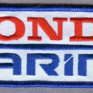 Puede incluir: Un parche rectangular bordado con fondo blanco y borde azul. El texto HONDA está bordado en letras mayúsculas rojas sobre una línea horizontal azul, con la palabra MARINE bordada en letras mayúsculas azules debajo de la línea.