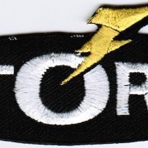 以下が含まれることがあります： 黒い背景に白い文字でSTORMと刺繍された楕円形のワッペン。文字Oの上には金色の稲妻のアイコンが配置されており、黒い楕円形の縁取りの上端からわずかに突き出ている。