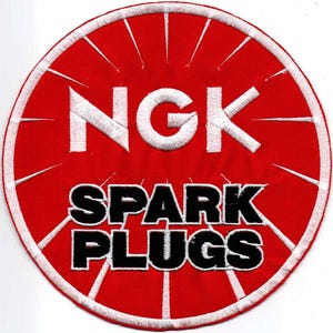 May include: A circular embroidered patch featuring a vibrant red background with white stitched radial lines. The text NGK is embroidered in large white letters at the top, while the words SPARK PLUGS appear in bold black letters below.