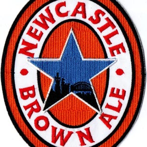 May include: An oval embroidered patch featuring a bright orange background with a white border. The text NEWCASTLE BROWN ALE is stitched in red. A blue star is centred, containing a black silhouette of a city skyline and a bridge.
