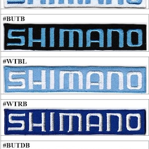 Peut inclure: Une collection de cinq écussons rectangulaires brodés présentant le mot SHIMANO en lettres capitales. Les écussons affichent diverses combinaisons de couleurs, notamment du texte bleu clair sur fond blanc, noir, bleu ciel, bleu roi et bleu marine foncé.