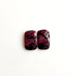 May include: Two rectangular gemstones with rounded corners. The stones are a deep burgundy colour with black inclusions, set against a white background. The gems have a faceted surface.
