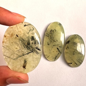 Puede incluir: Tres piedras preciosas ovaladas pulidas. Las piedras son de color amarillo verdoso pálido con inclusiones negras. La piedra más grande está sujeta por un dedo. Las piedras preciosas son lisas y translúcidas.