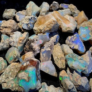 Può includere: Una collezione di pietre di opale grezze e non lucidate con forme irregolari e texture ruvide e terrose. Le pietre mostrano bagliori iridescenti di luce verde, blu e arancione su una base lattiginosa, traslucida e bianco-grigiastra.