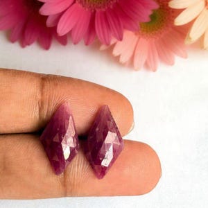 Puede incluir: Dos piedras preciosas sueltas talladas en forma de diamante con un tono magenta y púrpura intenso. Las piedras presentan una superficie geométrica multifacetada que refleja la luz, mostrando un borde superior e inferior afilado y un acabado pulido.
