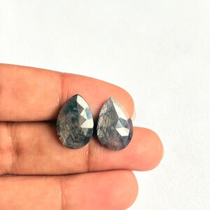 May include: Two faceted, teardrop-shaped gemstones with a dark blue-gray color. The gems have a complex, multi-faceted cut, reflecting light. The stones are held against a white background.