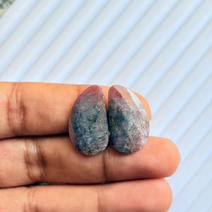 Puede incluir: Dos piedras preciosas ovaladas con una mezcla de tonos rojos, grises y azules. Las piedras tienen un aspecto pulido y facetado, con inclusiones visibles. Las piedras preciosas se muestran sobre un fondo blanco.