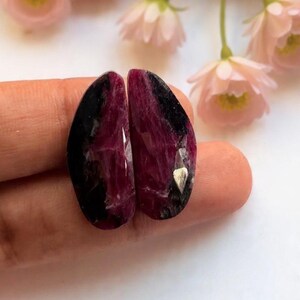Könnte beinhalten: Zwei polierte Edelsteine, jeweils mit einem tiefen Burgunder- und Schwarzfarbverlauf. Die Steine haben eine Tropfenform und eine reflektierende Oberfläche. Rosa Blumen sind im Hintergrund.