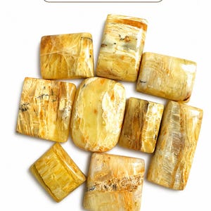 May include: A collection of polished, rectangular and oval-shaped gemstones in shades of yellow, brown, and cream. The stones have a natural, marbled appearance. The image includes the text "ZUGemsShop" at the top.