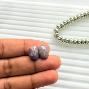 Op de afbeelding: Twee ovale, gefacetteerde edelstenen in tinten lavendel en grijs worden getoond. De edelstenen worden in een hand gehouden, met een zilveren kralenketting op de achtergrond. De edelstenen worden waarschijnlijk gebruikt voor het maken van sieraden.