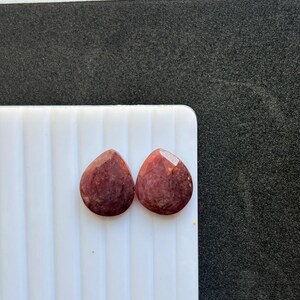 Puede incluir: Dos gemas facetadas en forma de lágrima con un tono marrón rojizo intenso. Las gemas se exhiben sobre una superficie blanca y acanalada, sobre un fondo oscuro. Las piedras tienen una superficie pulida y reflectante.