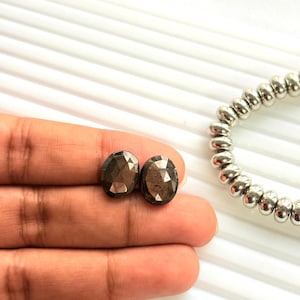 Könnte beinhalten: Zwei ovale, facettierte Edelsteine in Braun- und Grautönen werden auf einer Hand präsentiert. Im Hintergrund ist ein silbernes Perlenarmband zu sehen. Die Edelsteine sind wahrscheinlich für die Schmuckherstellung bestimmt.