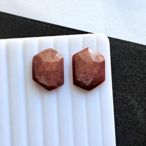 May include: Two faceted, reddish-brown gemstone cabochons with a hexagonal shape. The gemstones are displayed on a white, ridged surface, with a black and white background. The gemstones have a polished, reflective surface.