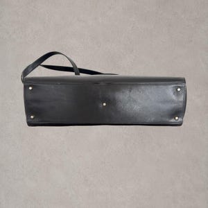 Puede incluir: Un bolso de mano de cuero negro visto desde la parte inferior, con una base rectangular que presenta cuatro peque&ntilde;os pies met&aacute;licos circulares en las esquinas y un pie met&aacute;lico central, junto con una correa de hombro de cuero negro lateral.