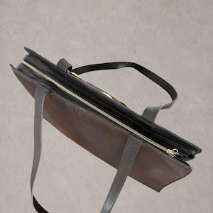 Puede incluir: Un bolso tote de cuero marr&oacute;n oscuro con cierre de cremallera superior y tirador dorado. El bolso incluye dos correas de hombro de cuero negro unidas al borde superior y un detalle decorativo dorado en el panel exterior frontal.