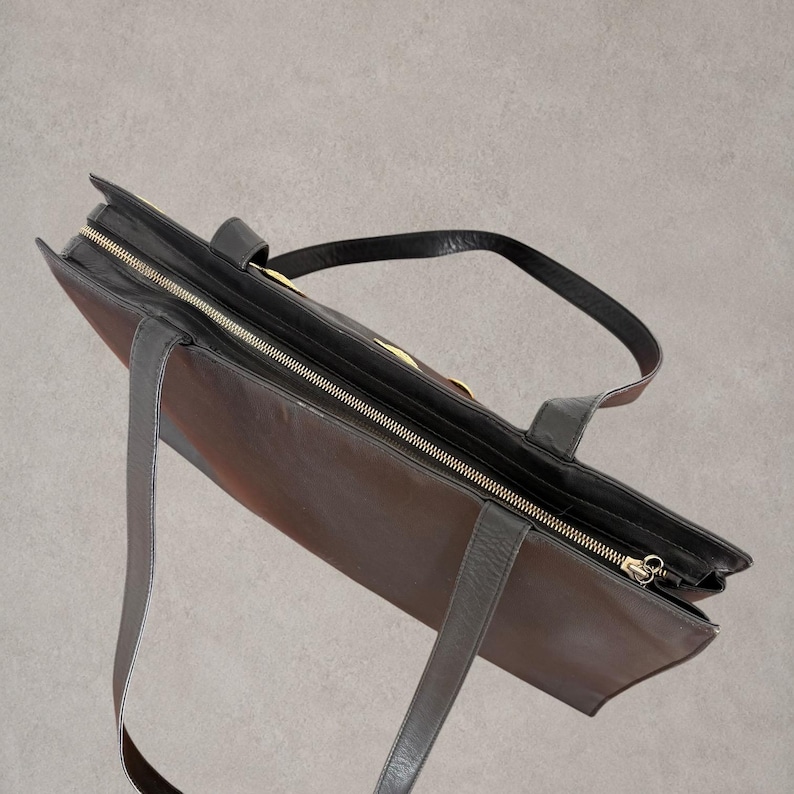 Puede incluir: Un bolso tote de cuero marr&oacute;n oscuro con cierre de cremallera superior y tirador dorado. El bolso incluye dos correas de hombro de cuero negro unidas al borde superior y un detalle decorativo dorado en el panel exterior frontal.
