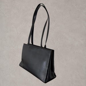 Puede incluir: Un bolso tote rectangular de color negro s&oacute;lido con acabado liso, base estructurada y dos correas de hombro largas y delgadas unidas al borde superior. El bolso presenta una silueta limpia con una amplia abertura y fuelles laterales.