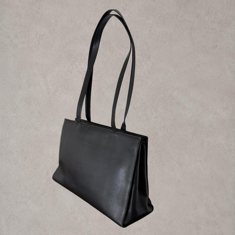 Puede incluir: Un bolso tote rectangular de color negro s&oacute;lido con acabado liso, base estructurada y dos correas de hombro largas y delgadas unidas al borde superior. El bolso presenta una silueta limpia con una amplia abertura y fuelles laterales.